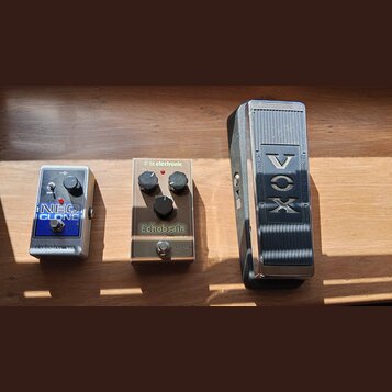 Na dřevěném povrchu leží tři kousky vybavení. Vlevo je stříbrný efektový pedál s nápisy „NEO CLONE“ a „electro-harmonix“. Uprostřed je stříbrný pedál…