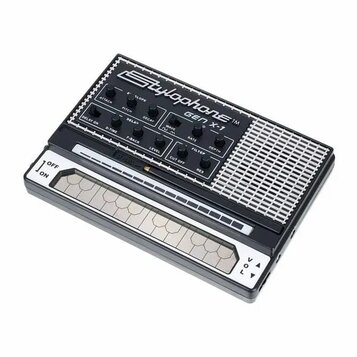 Černý přenosný syntetizátor s bílým nápisem Stylophone TM GEN X-1. Horní panel obsahuje řadu ovládacích knoflíků s popisky jako ATTACK, PITCH, DECAY,…