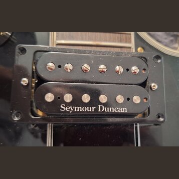 Fotografie zachycuje dva elektrické snímače typu humbucker v černém plastovém rámečku. Spodní snímač nese bílý nápis Seymour Duncan a má viditelné šr…