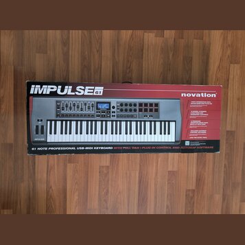 Fotografie znázorňuje krabici s MIDI klaviaturou v barvě stříbrné a černé. Na krabici jsou nápisy IMPULSE 61, novation a text 61 NOTE PROFESSIONAL US…