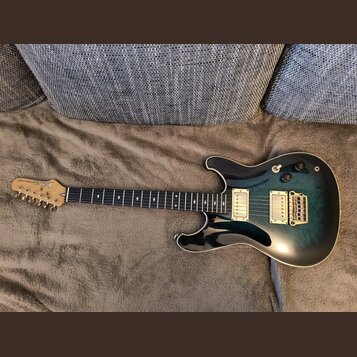 Elektrická kytara s modro-černým přechodovým lakem a krčkem z tmavého dřeva. Na hlavě je viditelné logo Ibanez a nápis RG SERIES. Nástroj má dva humb…