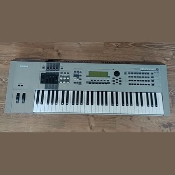 Šedý digitální klavír s bílými a černými klávesami. Na levé straně je nápis YAMAHA a vpravo nápis MOTIF 6. Přístroj obsahuje displej, několik posuvní…