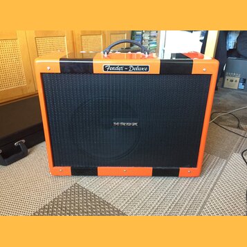 Kytarový kombozesilovač s oranžovým povrchem a černými vertikálními pruhy. Na horní hraně je stříbrný štítek s nápisem Fender - Deluxe a uprostřed če…