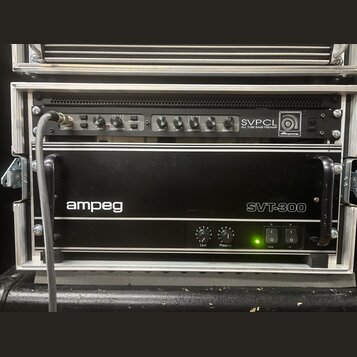 V hliníkovém racku jsou umístěna dvě černá zařízení značky Ampeg. Horní jednotka nese nápis SVPCL ALL TUBE BASS PREAMP a má řadu ovládacích knoflíků…