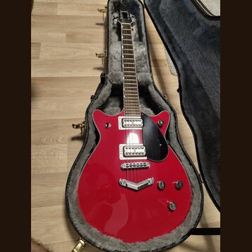 Červená elektrická kytara s černým pickguardem v otevřeném kufru s šedou vnitřní výstuhou. Na hlavě hmatníku je bílý nápis Gretsch. Kytara má dva hum…