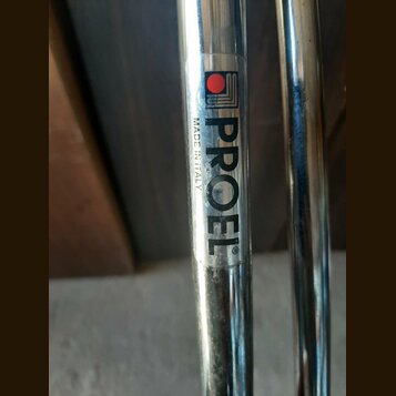 Detail chromovaného kovového trubkového nástroje. Na povrchu je nalepený štítek s černým nápisem PROEL® a menším svislým textem MADE IN ITALY. Nad ná…