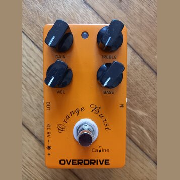 Oranžový efekt pedál s nápisy Orange Burst, Cadine a OVERDRIVE. Pedál má čtyři černé ovládací knoflíky s popisky GAIN, TREBLE, VOL a BASS. V dolní čá…