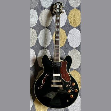 Černá polodublá elektrická kytara s bílým lemováním a hnědým hmatníkem. Na hlavě je bílý nápis Epiphone a dekorativní ornament. Tělo má dva humbucker…