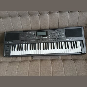 Černý digitální klavír s bílými a černými klávesami. Na levé straně je bílý nápis Roland a vpravo nápis EXR-5s INTERACTIVE ARRANGER. Horní panel obsa…