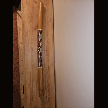 Dřevěný didgeridoo v přirozené hnědé barvě. Střed nástroje je zdobený ornamentálním vzorem v červených, bílých a žlutých odstínech. Na nástroji nejso…