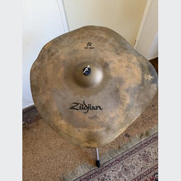Kovový činel s nepravidelným, vlnitým okrajem a matným, patinovaným povrchem. Na povrchu jsou čitelné nápisy „fx RAW CRASH“ a logo „Zildjian“. V prav…