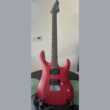 Červená elektrická kytara s matným povrchem a tmavým hnedým hmatníkem. Na hlavě je bílý nápis Cort. Nástroj má dva černá humbuckery, jeden ovládací k…