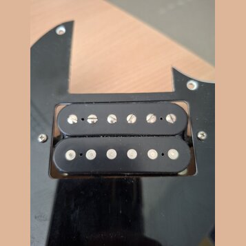 Fotografie detailně zobrazuje černý humbucker snímač s dvěmi řadami magnetických pólů osazených šrouby a kolíky. Snímač je umístěn v černém plastovém…