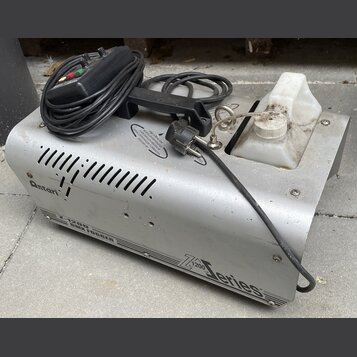 Stříbrný kouřový stroj z kovu s bílou plastovou nádržou na tekutinu. Na přístroji jsou nápisy „Antari“, „Z-1200 SMOKE FOGGER“ a „Z 1200 series“. Povr…
