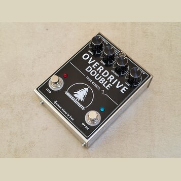 Kytarový efekt pedál v kovovém stříbrném šasi s černým horním panelem. Na panelu je bílý nápis OVERDRIVE DOUBLE, TRUE BYPASS a logo znázornující jehl…