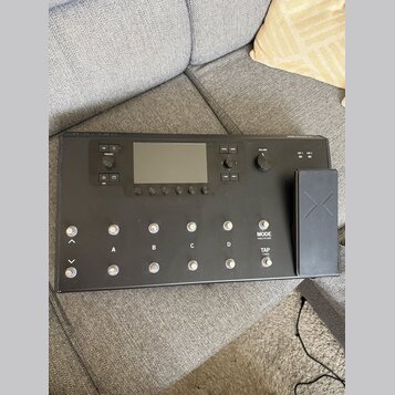 Černý kytarový procesor s displejem a ovládacími prvky. Na panelu jsou nápisy PRESET, VIEW, VOLUME, MODE HOLD TO EDIT a TAP TUNER, dále označení tlač…