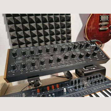 Černý analogový syntetizátor v dřevěném rámu s množstvím ovládacích knoflíků a přepínačů. V pravém horním rohu je bílé logo Korg. Na panelu jsou čite…