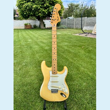 Elektrická kytara s žlutým trupem, bílým pickguardem a hnedým javorovým hmatníkem. Na hlavě je vidět logo Fender a nápis STRATOCASTER. Nástroj má tři…