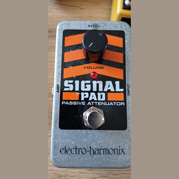 Kytarový pedál v stříbrném kovovém provedení s černým a oranžovým panelem. Na panelu je nápis SIGNAL PAD PASSIVE ATTENUATOR, označení VOLUME u ovláda…