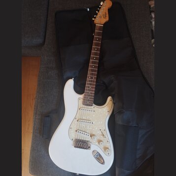 Bílá elektrická kytara s perleťovým pickguardem a hnedým hmatníkem. Na hlavě je nápis Squier STRAT. Nástroj leží na černém softcasu. Povrch kytary vy…