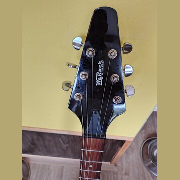 Černá hlava elektrické kytary s chromovanými ladičkami. Uprostřed je bílý nápis „Werner GUITARS“. Povrch lakovaného dřeva vykazuje drobné oděrky a zn…