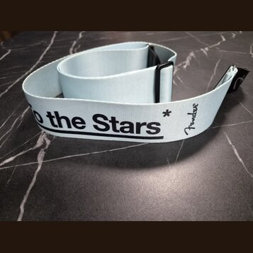 Světle modrý textilní popruh k hudebnímu nástroji s černými plastovými prvky. Na popruhu je černý nápis „to the Stars“ a logo „Fender“. Povrch popruh…