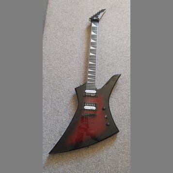 Elektrická kytara s ostře hranatým korpusem v černo-červeném přechodovém lakování. Na hlavě hmatníku je bílý nápis Cort. Nástroj má dva humbuckery, d…