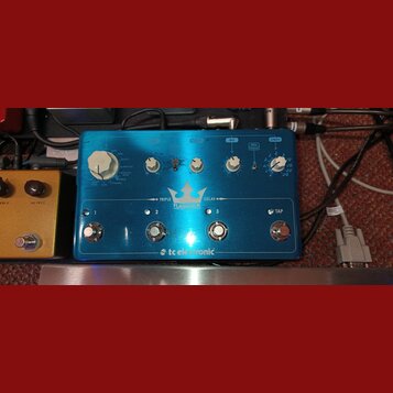 Modrý efekt pedál s nápisy TRIPLE FLASHBACK a tc electronic. Na horní části jsou čtyři bílé otočné ovladače a přepínače s popisky TAPE, TUBE, SPACE,…