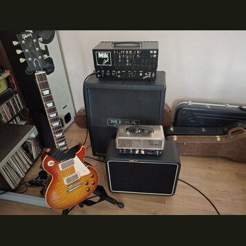 Elektrická kytara s trupem v barevném přechodu od žluté po červenou a bílým pickguardem. Na hlavě kytary je nápis Gibson a La Paul. Vedle ní stojí če…