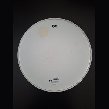 Bílá kruhová bubenová koží s bílým plastovým okrajem. V horní části je černý nápis 15" a v dolní části logo s korunkou a texty REMO, UT DRUM HEAD, MA…