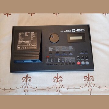 Černý digitální MIDI sekvencer s nápisem KAWAI Q-80 DIGITAL MIDI SEQUENCER. Zařízení má malý displej, ovládací tlačítka a vlevo slot pro diskety. Pov…