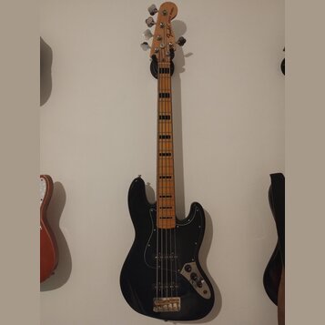 Černá elektrická basová kytara s černým pickguardem a hnedým hmatníkem. Na hlavě je viditelný nápis Fender JAZZ BASS. Nástroj vykazuje běžné známky p…