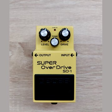 Žlutý efekt pedál s nápisy SUPER OverDrive a SD-1. Na horní části jsou tři stříbrné ovládací knoflíky s popisky LEVEL, TONE a DRIVE a červená kontrol…