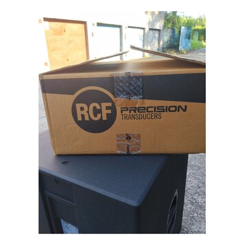 Kartonová krabice s nápisy RCF PRECISION TRANSDUCERS položená na černém reproduktorovém boxu. Krabice je z hnědého kartonu s černým pruhem a je slepe…
