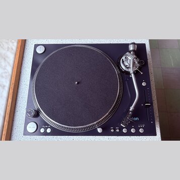 Černý gramofon s kulatým talířem potaženým tmavě šedou plstí a stříbrným vlnitým okrajem. Na raménku je viditelné logo "aton" s modrým symbolem. Ovlá…