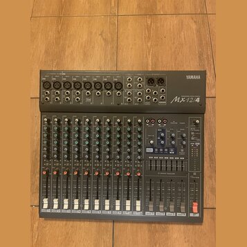 Šedý mixovací pult značky YAMAHA, model Mx12/4. Horní panel obsahuje vstupy, konektory a ovládací prvky s nápisy jako INPUT, GAIN, HIGH, LOW, MON a E…
