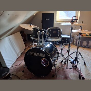 Černá bicí souprava značky YAMAHA s chromovaným hardwarem. Na basovém bubně je bílý nápis YAMAHA s logem a v dolní části malý kulatý štít. V pozadí s…