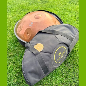 Na fotografii je kovový nástroj typu handpan v měděné barvě, umístěný v černé přepravce. Na povrchu nástroje je nápis „Handpan Design Sela“. Před nás…