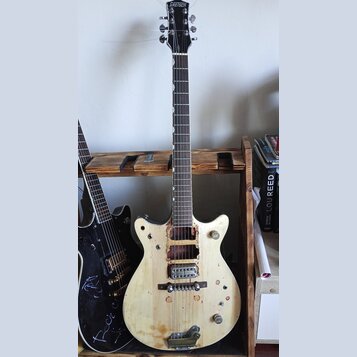 Elektrická kytara s bílým nebo vybledlým povrchem těla a tmavým hmatníkem. Na hlavě je nápis GRETSCH. Nástroj je nekompletní, chybí mu snímače a ovlá…