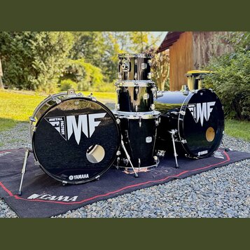 Černá bicí souprava složená z dvou basových bubnů, jednoho tom-tomu a jednoho floor tomu. Na předních blanách basových bubnů je bílý nápis METAL FACT…