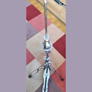 Stříbrný kovový pedál na hi-hat značky MAPEX. Nástroj stojí na vícebarevném koberci a je vybaven kovovou tyčí a mechanismem s utahovacími šrouby. Pov…