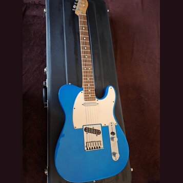 Modrá elektrická kytara typu Telecaster s bílým pickguardem a hnedým hmatníkem. Nástroj leží v černém tvrdém kufru. Na hlavě kytary je viditelné logo…