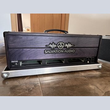 Černý transportní kufřík s dřevěným předním panelem a hliníkovým rámem. Na panelu je bílé logo s nápisem SALVATION AUDIO a grafickým symbolem vlny. V…