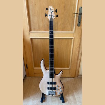 Elektrická basová kytara s přírodním dřevěným povrchem těla a hmatníkem z tmavého dřeva. Na hlavě je viditelné logo "Ibanez". Nástroj má dva humbucke…