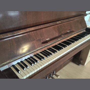 Tmavě hnědé dřevěné piano s klávesnicí. Povrch dřeva vykazuje známky opotřebení a drobné škrábance. Na klávesnici jsou vidět nečistoty a mírné změny…