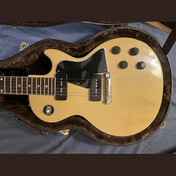 Světle žlutá elektrická kytara s černým pickguardem a čtyřmi ovládacími knoflíky v hnědém plišovém pouzdře. Nástroj má dva černávé humbuckery a stříb…
