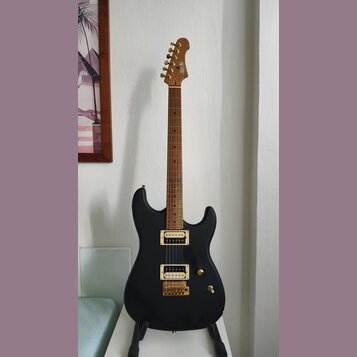 Černá elektrická kytara s matným povrchem a zlatým hardwarem. Na hlavě hmatníku je viditelné logo "Cort". Nástroj má dva humbuckery a zlatý most. Kyt…