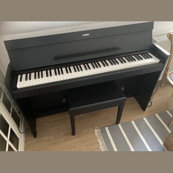 Černý digitální piano s klaviaturou a přívodním kabelem. Na horní části korpusu je bílé logo YAMAHA. Před nástrojem stojí černá pianová stolička. Pov…