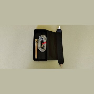 Černý úzký kufr, ve kterém je umístěn stříbrný mikrofon a srolovaný šedý kabel. Kabel je svázaný červeným plastovým stahovákem. V pravé části je přip…
