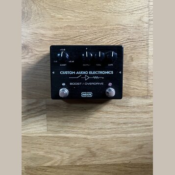 Černý efekt pedál s nápisy CUSTOM AUDIO ELECTRONICS a BOOST / OVERDRIVE. Na horní části jsou čtyři ovládací knoflíky s popisky BOOST (s rozsahem 0 dB…